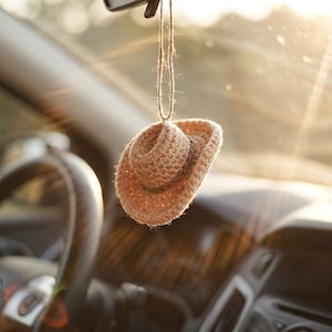 May include: A crocheted, tan-colored cowboy hat car charm hanging from a string. The hat is suspended in front of a car's dashboard and rearview mirror, with sunlight streaming in from the right. The hat has a classic cowboy hat shape.