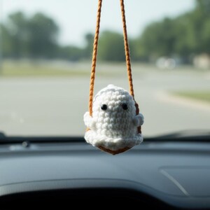 Peut inclure: Un porte-cl&eacute;s de voiture en forme de fant&ocirc;me blanc crochet&eacute;, avec des yeux noirs et une petite bouche, suspendu &agrave; une ficelle marron. Le porte-cl&eacute;s est suspendu devant un arri&egrave;re-plan flou de route et d'arbres.