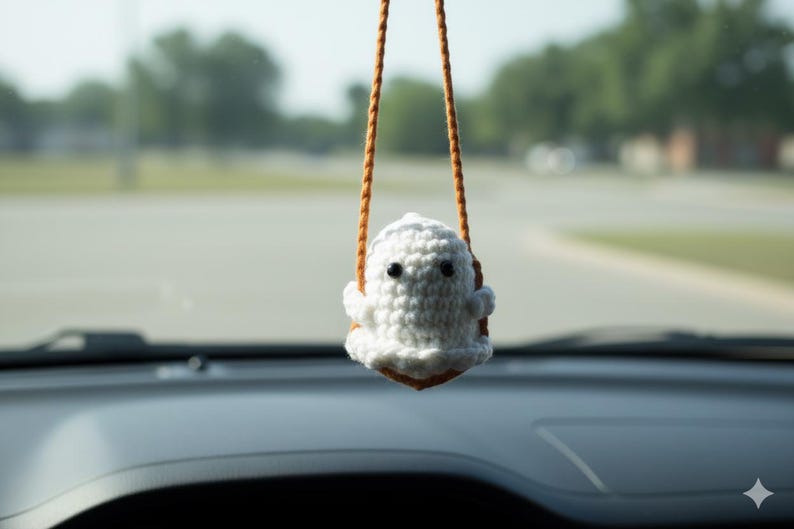 Peut inclure: Un porte-cl&eacute;s de voiture en forme de fant&ocirc;me blanc crochet&eacute;, avec des yeux noirs et une petite bouche, suspendu &agrave; une ficelle marron. Le porte-cl&eacute;s est suspendu devant un arri&egrave;re-plan flou de route et d'arbres.