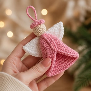 May include: A small crochet angel ornament featuring a pink dress, white wings, a beige head, and a pink hat with a matching pink loop at the top for hanging. The handcrafted textile figure is constructed with visible yarn stitches throughout.