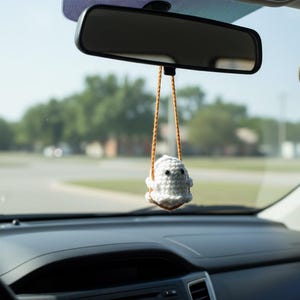 Peut inclure: Un porte-bonheur de voiture en forme de fant&ocirc;me crochet&eacute; blanc, suspendu au r&eacute;troviseur d'une voiture. Le fant&ocirc;me a des yeux noirs et est suspendu par des cordons orange-brun. Le tableau de bord et le pare-brise de la voiture sont visibles.