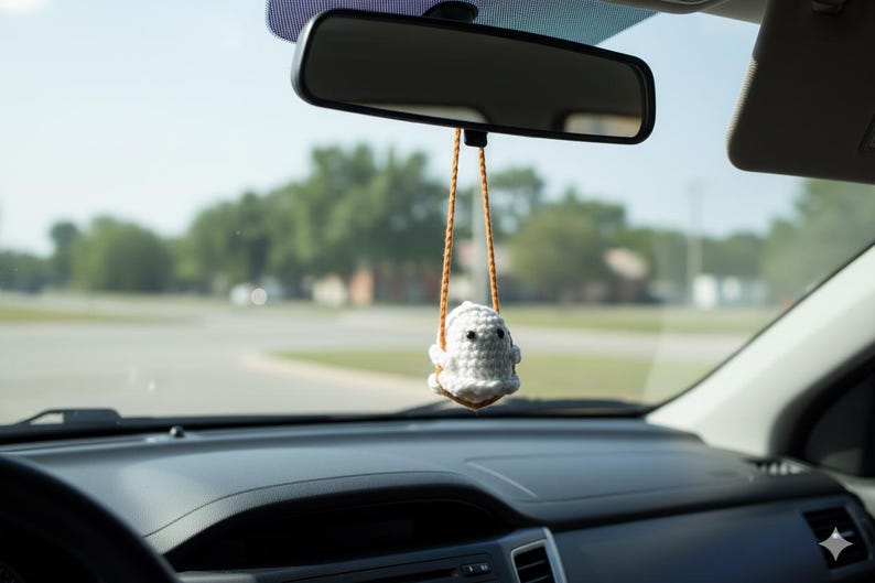 Peut inclure: Un porte-bonheur de voiture en forme de fant&ocirc;me crochet&eacute; blanc, suspendu au r&eacute;troviseur d'une voiture. Le fant&ocirc;me a des yeux noirs et est suspendu par des cordons orange-brun. Le tableau de bord et le pare-brise de la voiture sont visibles.