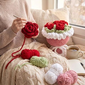 May include: A person crocheting a large red rose. A finished crochet flower arrangement in a pink pot with red roses, green leaves, and white trim sits nearby. Several balls of yarn in red, green, white, and pink are on a cream-colored blanket.