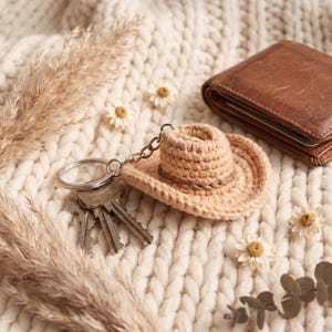 May include: A beige crocheted cowboy hat keychain with a silver chain and keyring, next to a set of keys and a brown leather wallet. The hat has a brown band around the crown. The items rest on a cream-colored, textured surface with dried flowers.