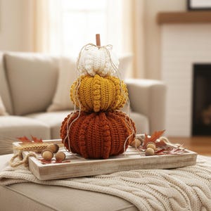 Puede incluir: Una pila decorativa de tres calabazas de ganchillo en colores oto&ntilde;ales: blanco, amarillo y naranja. Las calabazas est&aacute;n dispuestas sobre una bandeja de madera con decoraciones tem&aacute;ticas de oto&ntilde;o, incluyendo bellotas y hojas. Un art&iacute;culo r&uacute;stico de decoraci&oacute;n oto&ntilde;al para el hogar.