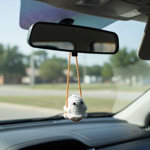 Peut inclure: Un porte-cl&eacute;s de voiture en forme de fant&ocirc;me crochet&eacute; blanc suspendu au r&eacute;troviseur d'une voiture. Le fant&ocirc;me a des yeux noirs et une petite bouche. Le charme est suspendu par un cordon marron et orange. L'int&eacute;rieur de la voiture est visible.