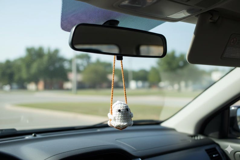 Peut inclure: Un porte-cl&eacute;s de voiture en forme de fant&ocirc;me crochet&eacute; blanc suspendu au r&eacute;troviseur d'une voiture. Le fant&ocirc;me a des yeux noirs et une petite bouche. Le charme est suspendu par un cordon marron et orange. L'int&eacute;rieur de la voiture est visible.