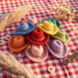 May include: A collection of colorful crochet cowboy hat keychains in various colors, including blue, orange, red, pink, yellow, and green. Each hat has a brown band and a silver keychain. The hats are arranged on a red and white checkered surface.