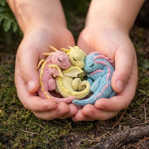 May include: Three crocheted baby dragons in pink, yellow, and blue are nestled in cupped hands. The dragons have closed eyes and are curled up together. The background is a natural setting with moss and foliage.