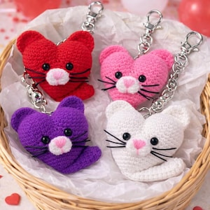 May include: Four crochet cat keyrings in red, pink, purple, and white, each with a heart-shaped head and silver keyring hardware. The cats have black whiskers and pink noses. The keyrings are in a woven basket with paper filling.