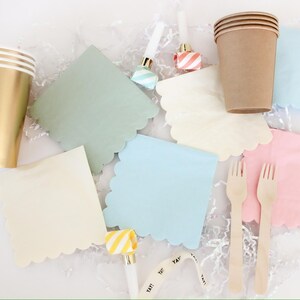 Op de afbeelding: Feestartikelen, waaronder gouden en bruine papieren bekers, pastelkleurige servetten met geschulpte randen in groen, blauw, roze en cr&egrave;me, houten vorken en toeters. Het woord "YAY!" staat op een feestartikel.