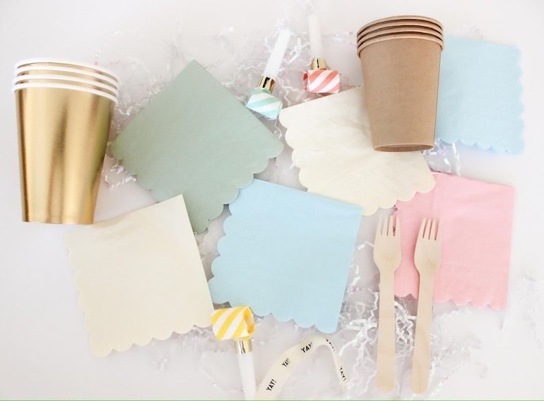 Op de afbeelding: Feestartikelen, waaronder gouden en bruine papieren bekers, pastelkleurige servetten met geschulpte randen in groen, blauw, roze en cr&egrave;me, houten vorken en toeters. Het woord "YAY!" staat op een feestartikel.