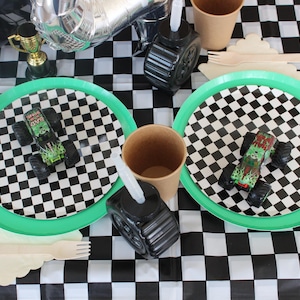 Op de afbeelding: Een feesttafel met een zwart-wit geblokt tafelkleed. Twee borden met groene rand en monstertruck speelgoed staan op de tafel. Andere items zijn papieren bekers, houten bestek en een zwarte bandvormige container.