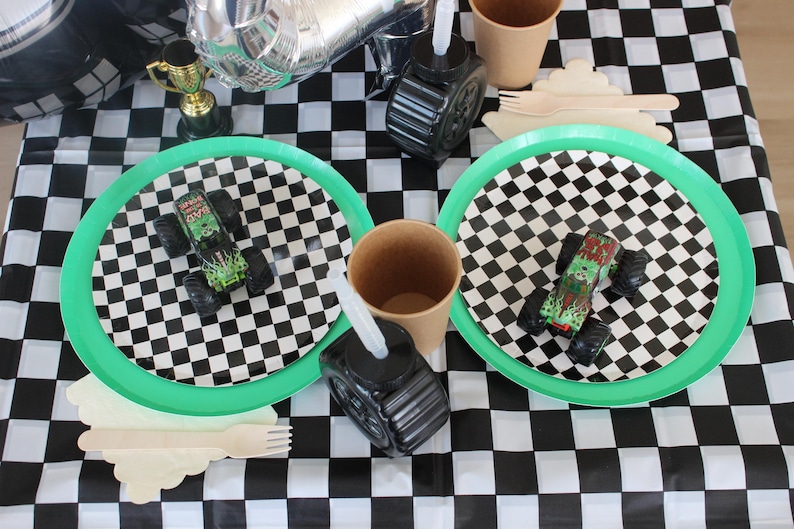 Op de afbeelding: Een feesttafel met een zwart-wit geblokt tafelkleed. Twee borden met groene rand en monstertruck speelgoed staan op de tafel. Andere items zijn papieren bekers, houten bestek en een zwarte bandvormige container.