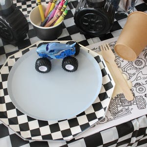 Op de afbeelding: Een feesttafel met een blauw monstertruckspeelgoed op een lichtblauw bord. Inclusief een zwart-wit geblokt tafelkleed, papieren bekers, kleurpotloden en een kleurplaat met raceauto-ontwerpen.