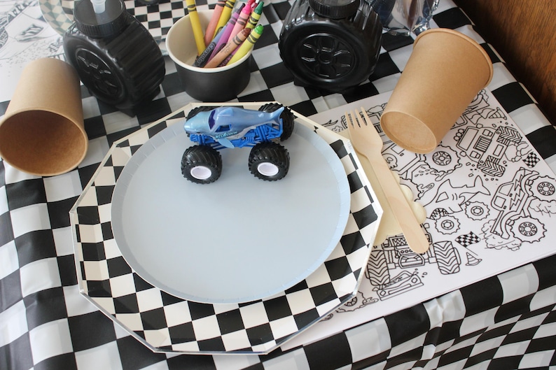 Op de afbeelding: Een feesttafel met een blauw monstertruckspeelgoed op een lichtblauw bord. Inclusief een zwart-wit geblokt tafelkleed, papieren bekers, kleurpotloden en een kleurplaat met raceauto-ontwerpen.