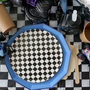 Op de afbeelding: Een feesttafelsetting met een zwart-wit geblokt thema. Inclusief een blauwe achthoekige plaat, een geblokte plaat, bruine papieren bekers, een houten vork en speelgoed monstertrucks. De tafel is bedekt met een zwart-wit geblokt tafelkleed.