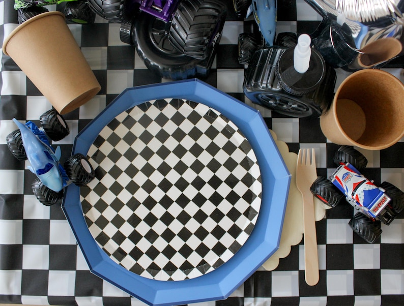 Op de afbeelding: Een feesttafelsetting met een zwart-wit geblokt thema. Inclusief een blauwe achthoekige plaat, een geblokte plaat, bruine papieren bekers, een houten vork en speelgoed monstertrucks. De tafel is bedekt met een zwart-wit geblokt tafelkleed.