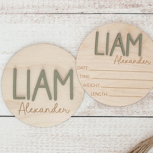 May include: Two round wooden baby announcement plaques. The name "LIAM" is in raised, olive green letters, with "Alexander" in script below. One plaque has fill-in-the-blank lines for date, time, weight, and length.