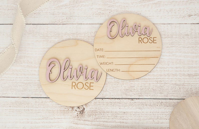May include: Two circular wooden baby announcement plaques. The name "Olivia Rose" is in pink, raised lettering. One plaque has lines for date, time, weight, and length. The plaques are on a light wood background.