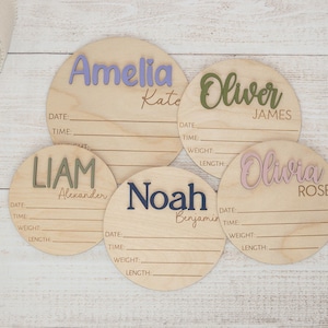 May include: Five wooden birth announcement signs with names in various colours: Amelia, Oliver, Liam, Noah, and Olivia. Each sign has lines for date, time, weight, and length. The names are in lavender, green, olive, navy, and pink.