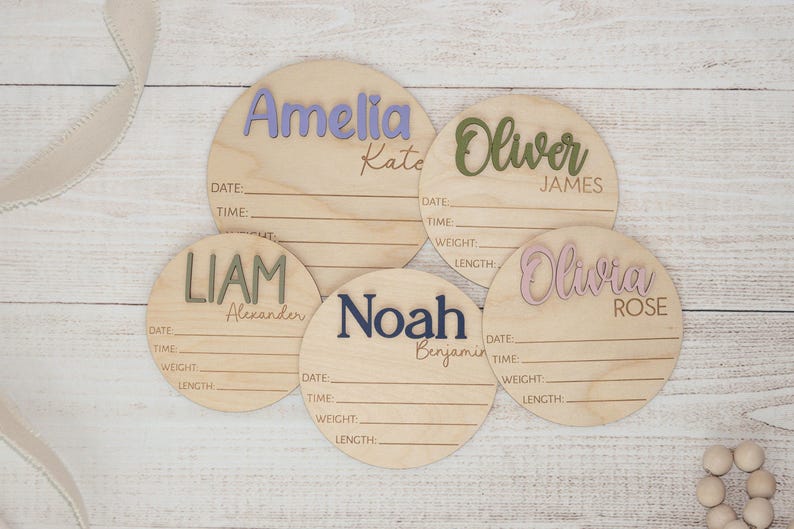 May include: Five wooden birth announcement signs with names in various colours: Amelia, Oliver, Liam, Noah, and Olivia. Each sign has lines for date, time, weight, and length. The names are in lavender, green, olive, navy, and pink.