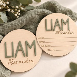 Puede incluir: Dos placas circulares de madera con el nombre LIAM Alexander en letras verde salvia en relieve. Una placa est&aacute; en blanco, mientras que la otra incluye l&iacute;neas grabadas con las etiquetas DATE, TIME, WEIGHT y LENGTH para registrar datos.