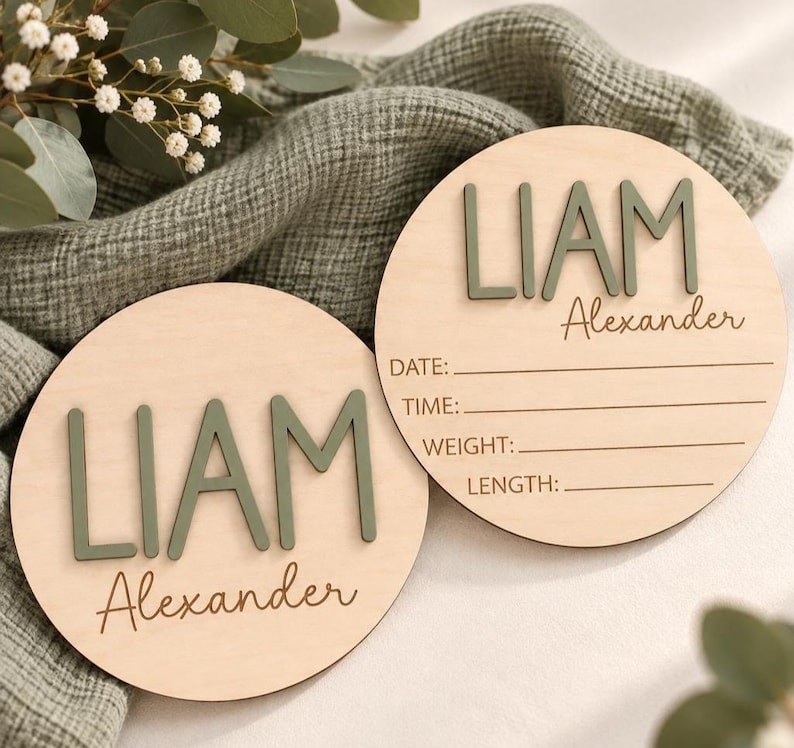 Puede incluir: Dos placas circulares de madera con el nombre LIAM Alexander en letras verde salvia en relieve. Una placa est&aacute; en blanco, mientras que la otra incluye l&iacute;neas grabadas con las etiquetas DATE, TIME, WEIGHT y LENGTH para registrar datos.