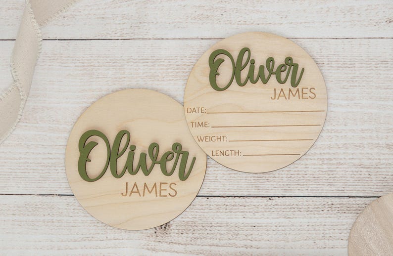May include: Two circular wooden plaques for a baby announcement. The name "Oliver James" is in green lettering. One plaque has lines for date, time, weight, and length. The background is a light wood grain.