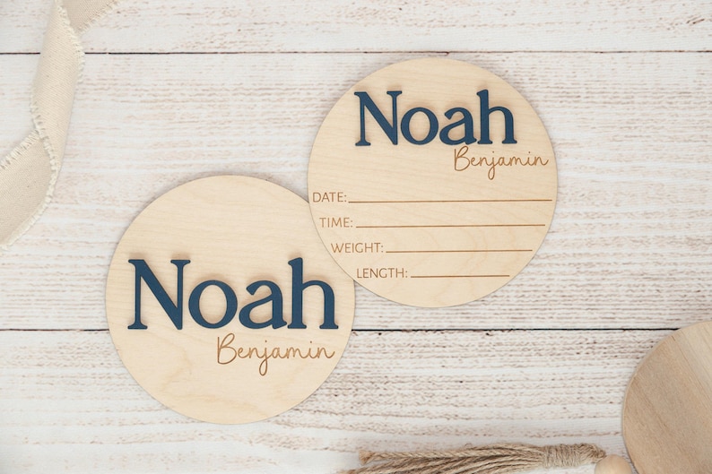 May include: Two round wooden baby announcement signs. The signs are light-coloured wood with the name "Noah" in navy blue, and "Benjamin" in a smaller, lighter font. One sign has lines for date, time, weight, and length.
