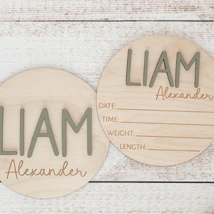 Puede incluir: Dos placas circulares de madera clara con el nombre LIAM Alexander en letras verde salvia en relieve. Una de las placas incluye l&iacute;neas grabadas con las etiquetas DATE, TIME, WEIGHT y LENGTH para registrar los datos de nacimiento.