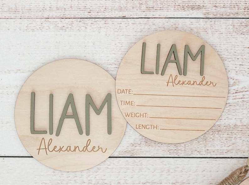 Puede incluir: Dos placas circulares de madera clara con el nombre LIAM Alexander en letras verde salvia en relieve. Una de las placas incluye l&iacute;neas grabadas con las etiquetas DATE, TIME, WEIGHT y LENGTH para registrar los datos de nacimiento.