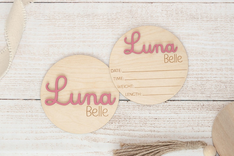 May include: Two circular wooden baby announcement plaques. One features the name "Luna Belle" in pink script. The other has "Luna Belle" and lines for date, time, weight, and length. Set against a light wood grain backdrop.