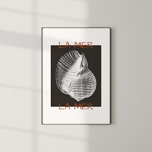 May include: A framed art print with a black and white seashell illustration on a black background. The words "LA MER" are printed in orange above and below the shell. The print is presented in a black frame, suitable for wall display.