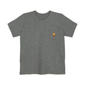 Puede incluir: Una camiseta gris jaspeada con un pequeño bolsillo en el pecho. El bolsillo presenta un gráfico amarillo y naranja de una piruleta. La camiseta tiene mangas cortas y cuello redondo.