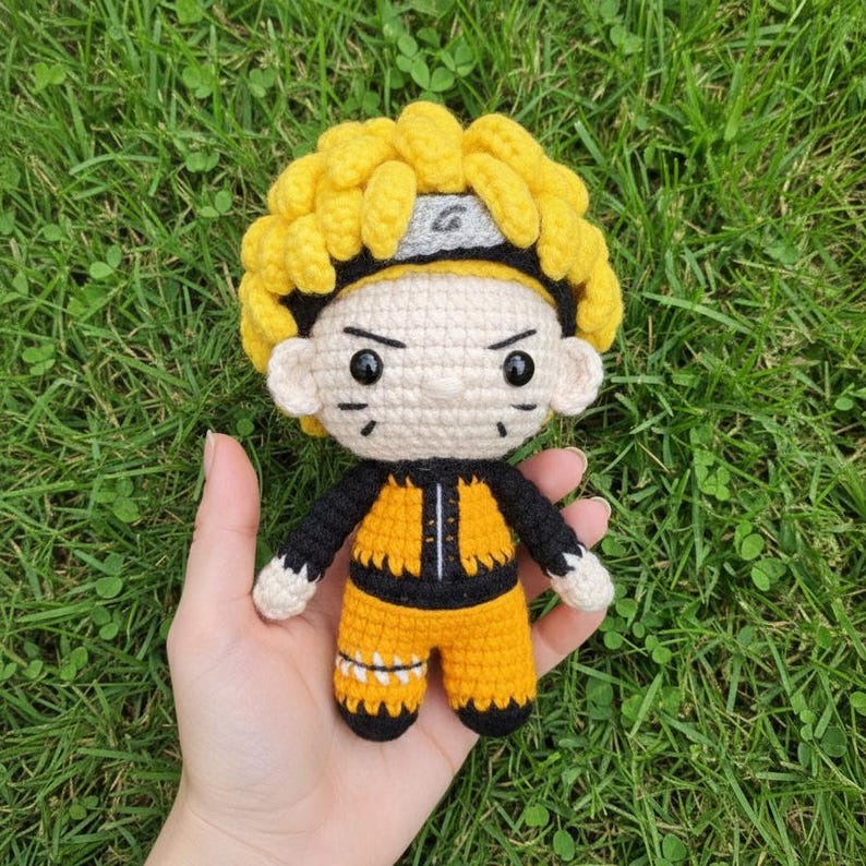 May include: A handmade crochet doll featuring a character with spiky yellow hair, a gray headband, and a black and orange outfit. The doll has black eyes and is held against a backdrop of green grass.