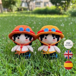 Puede incluir: Dos muñecos de ganchillo de un personaje que lleva un sombrero naranja con detalles azules, un borde rojo y una camisa amarilla. Los muñecos tienen el pelo negro y están de pie en hierba verde. Los muñecos llevan pantalones negros y azules. El fondo está borroso.