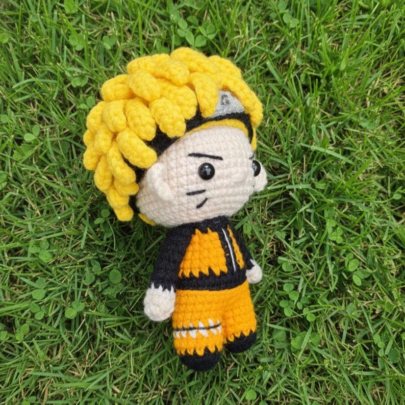 May include: A handmade crochet doll of a character with spiky yellow hair, a black headband, and an orange and black outfit. The doll has a cream-colored face with black eyes and a stitched mouth. The doll is standing on green grass.