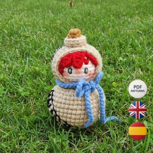 Könnte beinhalten: Handgemachte Häkelpuppe mit roten Haaren, einem blauen Schal und einem beigen Outfit. Die Puppe hat einen runden Kopf und eine kleine, braune Kappe. Die Puppe sitzt auf grünem Gras. Das Bild enthält auch einen Button mit dem Text "PDF PATTERNS" und Flaggen.