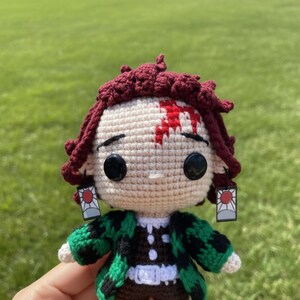 Pu&ograve; includere: Bambola all'uncinetto raffigurante un personaggio con capelli bordeaux, un segno rosso sulla fronte e occhi neri. La bambola indossa una giacca a fantasia verde e nera e orecchini con un disegno a sole rosso. Sfondo di erba verde.