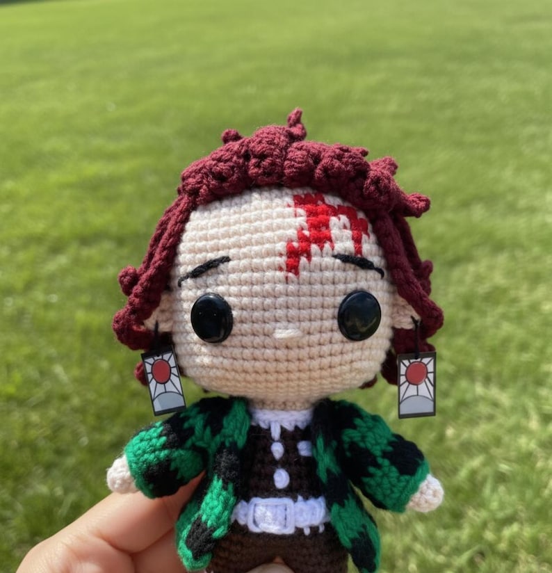Pu&ograve; includere: Bambola all'uncinetto raffigurante un personaggio con capelli bordeaux, un segno rosso sulla fronte e occhi neri. La bambola indossa una giacca a fantasia verde e nera e orecchini con un disegno a sole rosso. Sfondo di erba verde.