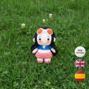 Puede incluir: Muñeca de ganchillo hecha a mano con pelo negro, gafas con montura naranja y un atuendo azul, blanco y rosa. La muñeca está sobre hierba verde. El patrón es PDF.