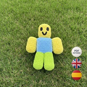 May include: A handmade, plush doll in the shape of a character with a yellow head, yellow arms, blue torso, and green legs. The doll has a smiling face and is made of a soft, textured material. The doll is lying on green grass.
