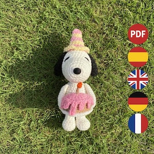 Peut inclure: Une figurine Snoopy au crochet portant un chapeau de fête et tenant un gâteau rose avec une bougie. La figurine est blanche avec des oreilles noires et un nez noir. Le gâteau a une base marron. L'arrière-plan est de l'herbe verte.