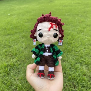 Pu&ograve; includere: Bambola all'uncinetto fatta a mano con un personaggio con capelli bordeaux, una cicatrice sulla fronte e abiti a fantasia neri e verdi. La bambola ha grandi occhi neri e piccoli orecchini. La bambola &egrave; tenuta in mano su uno sfondo verde.