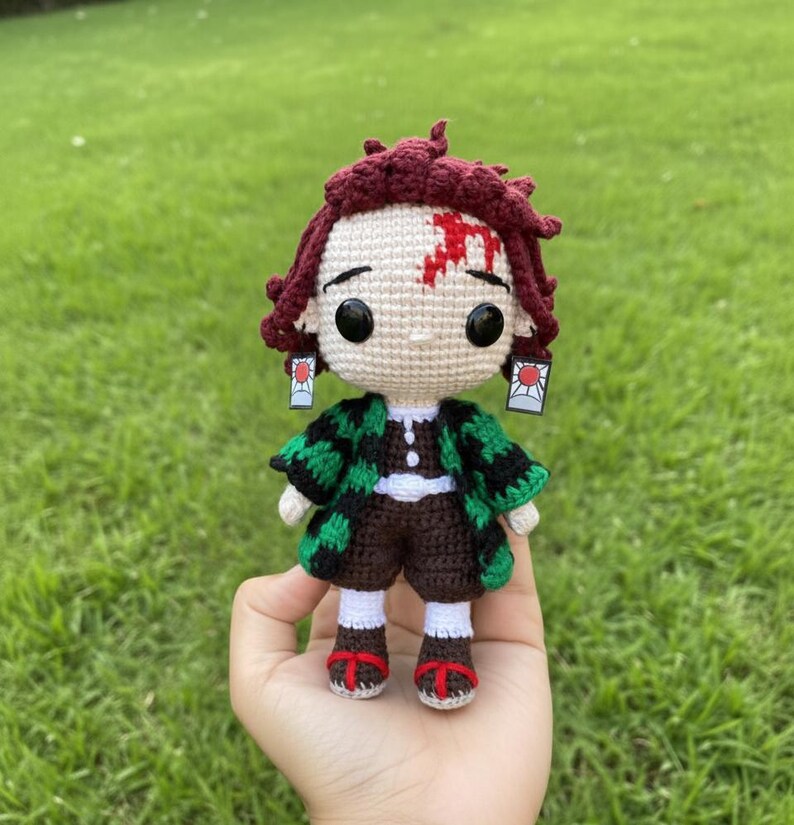 Pu&ograve; includere: Bambola all'uncinetto fatta a mano con un personaggio con capelli bordeaux, una cicatrice sulla fronte e abiti a fantasia neri e verdi. La bambola ha grandi occhi neri e piccoli orecchini. La bambola &egrave; tenuta in mano su uno sfondo verde.