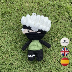 May include: A handmade crochet doll of a character with a black mask, black outfit, and a green shirt. The doll has white hair and a small loop at the top. The doll is on a bed of green grass. A white circle with the words "PDF PATTERNS" is in the background.