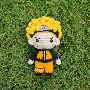 May include: A handmade crochet doll of a character with spiky yellow hair, a black and gray headband, and a determined expression. The doll wears an orange and black outfit. The doll is sitting on a bed of green grass.