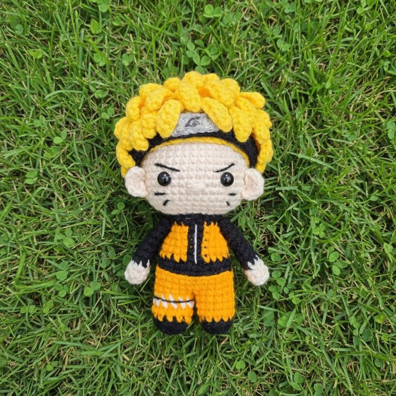 May include: A handmade crochet doll of a character with spiky yellow hair, a black and gray headband, and a determined expression. The doll wears an orange and black outfit. The doll is sitting on a bed of green grass.