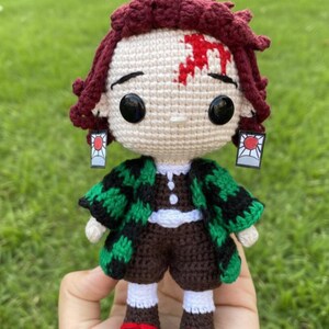 Pu&ograve; includere: Bambola all'uncinetto raffigurante un personaggio con capelli bordeaux, occhi neri e una cicatrice sulla fronte. La bambola indossa una giacca a fantasia verde e nera, pantaloncini marroni e scarpe con dettagli rossi. Sono presenti anche orecchini con un disegno a sole rosso.
